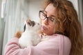 Attractive girl in pink clothes and eyeglasses with withe cat in her arms standing near the window at home. Pet owner Royalty Free Stock Photo