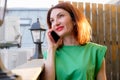 Attractive girl with red hair in a green blouse is talking on a mobile phone on the open terrace of a cafe Royalty Free Stock Photo