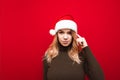 Attractive girl in a santa hat stands on a red background, looks into the camera and shows a gesture to think, touches her finger Royalty Free Stock Photo