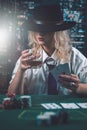 attractive girl in shirt and hat drinking whiskey and looking at poker cards in casino Royalty Free Stock Photo