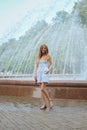 Attractive girl in short dress stands in front of fountain in summer park