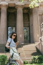 attractive girl in sunglasses sitting on bike and using smartphone near beautiful building with columns