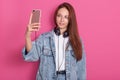 Attractive girl taking selfie against pink studio wall. Grinning woman in stylish denim jacket and basic whit t shirt, holding