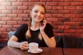 Attractive girl talking on phone while drinking coffee in cozy cafe against brick wall, smiling red haired lady spending free time Royalty Free Stock Photo