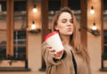 Attractive girl in warm clothes on a standing background of the building holds a paper cup with coffee in her hands, a kiss with Royalty Free Stock Photo