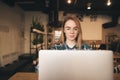 Attractive girl wearing glasses and a shirt uses a laptop in a cozy cafe, focuses on the screen and smiles. Teen girl with laptop Royalty Free Stock Photo