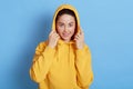 Attractive girl in yellow sweatshirt with hood looking at camera and smiling, stands against blue wall, keeps hands on her hood,