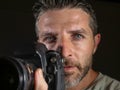 Attractive and handsome man on his 30d holding professional reflex photo camera next to his face isolated on black background in Royalty Free Stock Photo