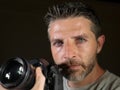 Attractive and handsome man on his 30d holding professional reflex photo camera next to his face isolated on black background in Royalty Free Stock Photo