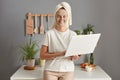 Attractive happy cheerful young woman posing in kitchen with towel on head standing with laptop in hands looking at camera working Royalty Free Stock Photo