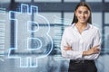 Attractive happy european businesswoman with creative bitcoin hologram standing on blurry office interior background.