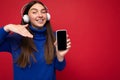 Attractive happy smiling young brunette woman wearing blue sweater isolated over red background holding and using mobile Royalty Free Stock Photo