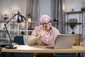 Attractive happy smiling young Muslim woman in hijab, sitting in headset in front of laptop on the desk, waving and Royalty Free Stock Photo