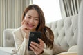 Attractive Asian female laying on sofa and chatting with her friends on her smartphone