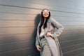 Attractive happy young hipster woman with positive smile in knitted hat in glasses in coat posing near gray building in the city. Royalty Free Stock Photo