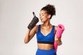 Attractive and healthy african-american female athlete with yoga mat, drinking water and smiling, standing against white Royalty Free Stock Photo