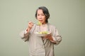 Healthy 60s aged-asian woman eating healthy green salad, standing isolated over green background Royalty Free Stock Photo