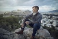 Attractive hipster with a beard is sitting on a rock high over Athens City with Acropolis view