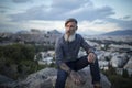 Attractive hipster with a beard is sitting on a rock high over Athens City with Acropolis view at sunset