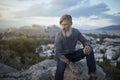 Attractive hipster with a grey beard is sitting on a rock high over Athens City with Acropolis view