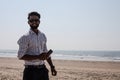 Attractive Indian man stands on beach , holding phone in his hands, smiling and looking at camera. Communications. Royalty Free Stock Photo