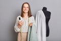 Attractive joyful cheerful professional female dressmaker with smartphone in hands standing isolated over gray background, Royalty Free Stock Photo