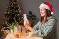 Attractive joyful woman dressed Santa cap and knitted shirt using smartphone scrolling online browsing web pages reading messages