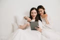 Attractive lesbian couple use mobile phone, tablet watch movie on bed. Beautiful sibling in pajamas lying down with blanket in Royalty Free Stock Photo