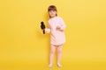 Attractive little girl with microphone wearing pink pale attire standing isolated over yellow background, charming vocalist sings