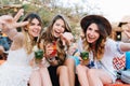 Attractive long-haired girls in stylish attires and accessories drink cocktails and posing with hands waving. Portrait