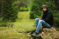 Attractive male cyclist with unshaven in a sports jacket, blue jeans, a buff and trekking boots sits on a tree trunk in the autumn