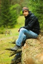 Attractive male cyclist with unshaven in a sports jacket, blue jeans, a buff and trekking boots sits on a tree trunk in the autumn