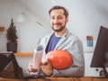 An attractive man with a beard smiles and poses for a photo with blueprints and a helmet sitting at a work table with computers.