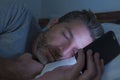 Attractive man falling asleep on bed late at night in dark light while networking on mobile phone or using online dating in