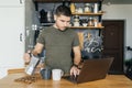 Attractive man uses a laptop and pours coffee in the interior of a home kitchen in the morning.
