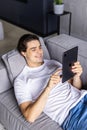 Attractive man using digital pad or tablet lying relaxed on couch at home connected to internet reading , watching the screen and Royalty Free Stock Photo