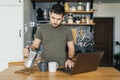 Attractive man working on a laptop and pouring coffee in the interior of a home kitchen Royalty Free Stock Photo