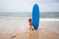 Attractive mature woman with curly hair, sunglasses and bikini, sitting clutching a blue surfboard on the shore of the beach. Royalty Free Stock Photo