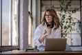 Attractive mid aged woman sitting at window and using her laptop for work Royalty Free Stock Photo