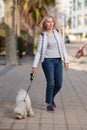 Attractive middle-aged blond woman walking with fluffy white dog in summer city