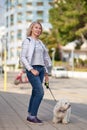 Attractive middle-aged blond woman walking with fluffy white dog in summer city