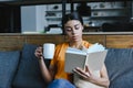 Attractive mixed race girl in orange shirt holding cup of tea and reading book on sofa Royalty Free Stock Photo