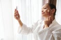 Attractive mixed race woman stands in front of sunlight by the window and looks at her reflection in the mirror while applying Royalty Free Stock Photo