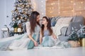 Attractive mom and her cute daughter in a gentle evening dresses sitting on the floor near the Christmas tree in a cozy