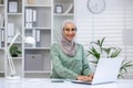 Attractive muslim woman wearing hijab and eyeglasses working on wireless laptop at desk in well-organized and bright Royalty Free Stock Photo