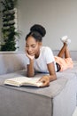 Attractive pensive African American girl lying on sofa dreamily reading book at home Royalty Free Stock Photo