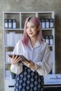 Attractive pink-haired Asian female employee standing in front of a desk Using a touch tablet computer, smiling, emailing business