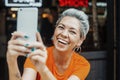 Attractive positive blonde woman in orange T-shirt making selfie at cafe