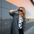 Attractive pretty young hipster woman in stylish clothes in fashionable black sunglasses poses in a city near a modern gray Royalty Free Stock Photo