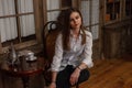 Attractive pretty young woman in a fashionable white shirt in black trendy jeans is sitting on wooden chair in the room. Stylish Royalty Free Stock Photo